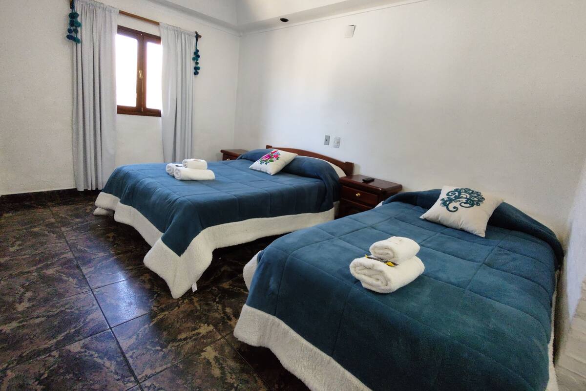 A hotel room with two beds, each with a blue bedspread and white pillows, and a window with curtains.