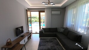 Ein modernes Wohnzimmer mit einem Ecksofa, einem Flachbildfernseher, einem Couchtisch aus Holz und einem großen Fenster mit durchsichtigen Vorhängen.