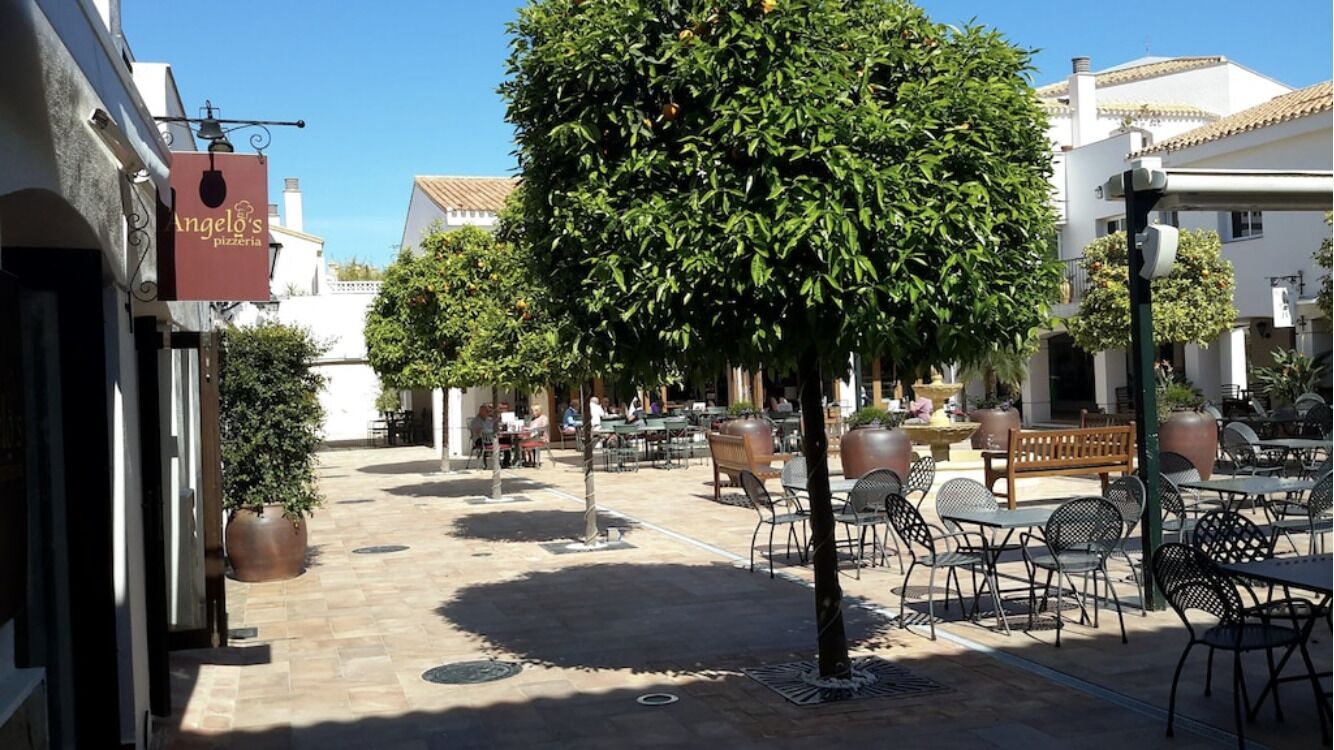 Un espace café en plein air avec des tables et des chaises, un arbre et une enseigne indiquant l'emplacement de la pizzeria d'Angelo.