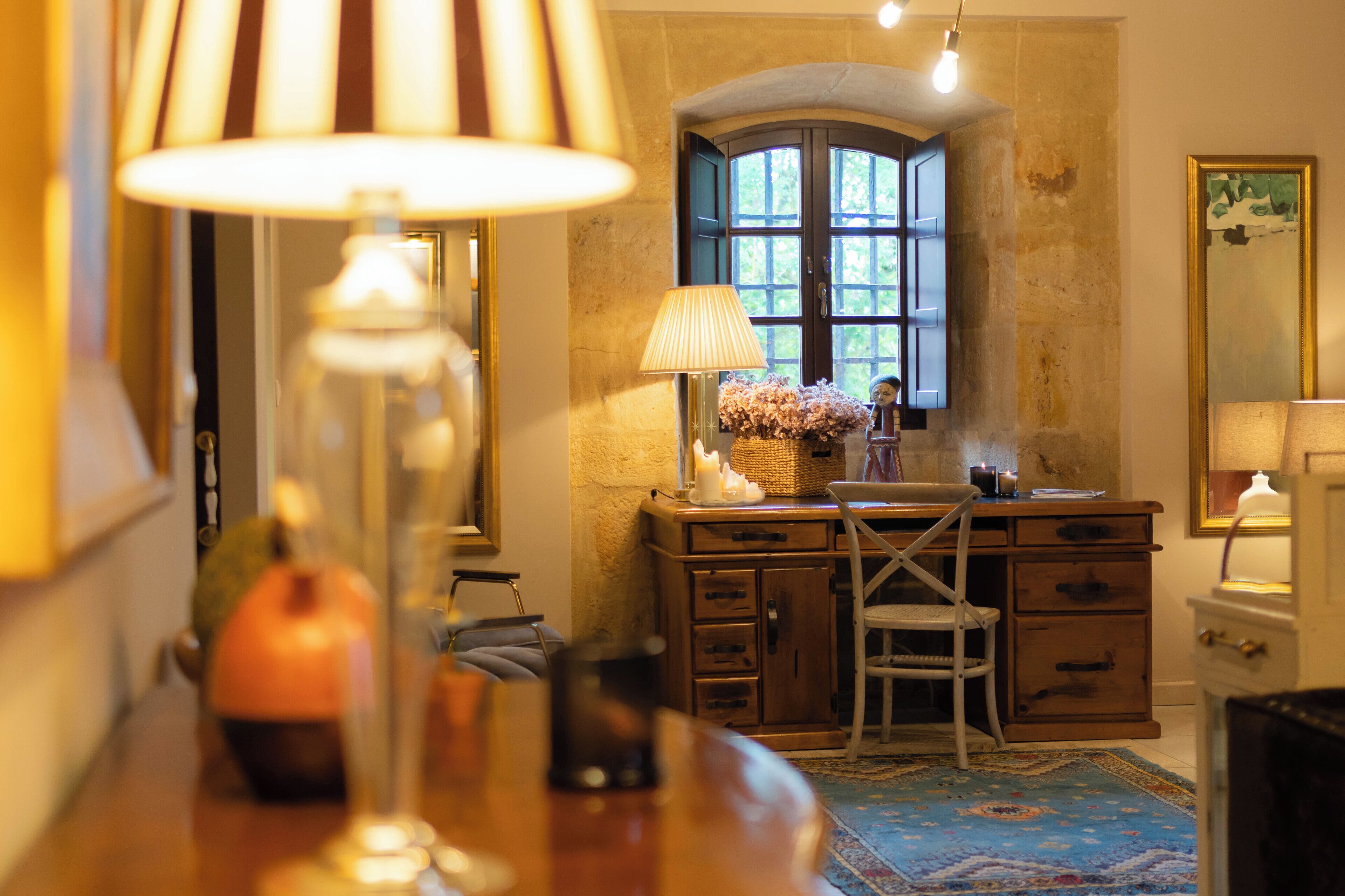 A room with a wooden desk, a chair, a lamp, a vase with flowers, and a window with a view of greenery.