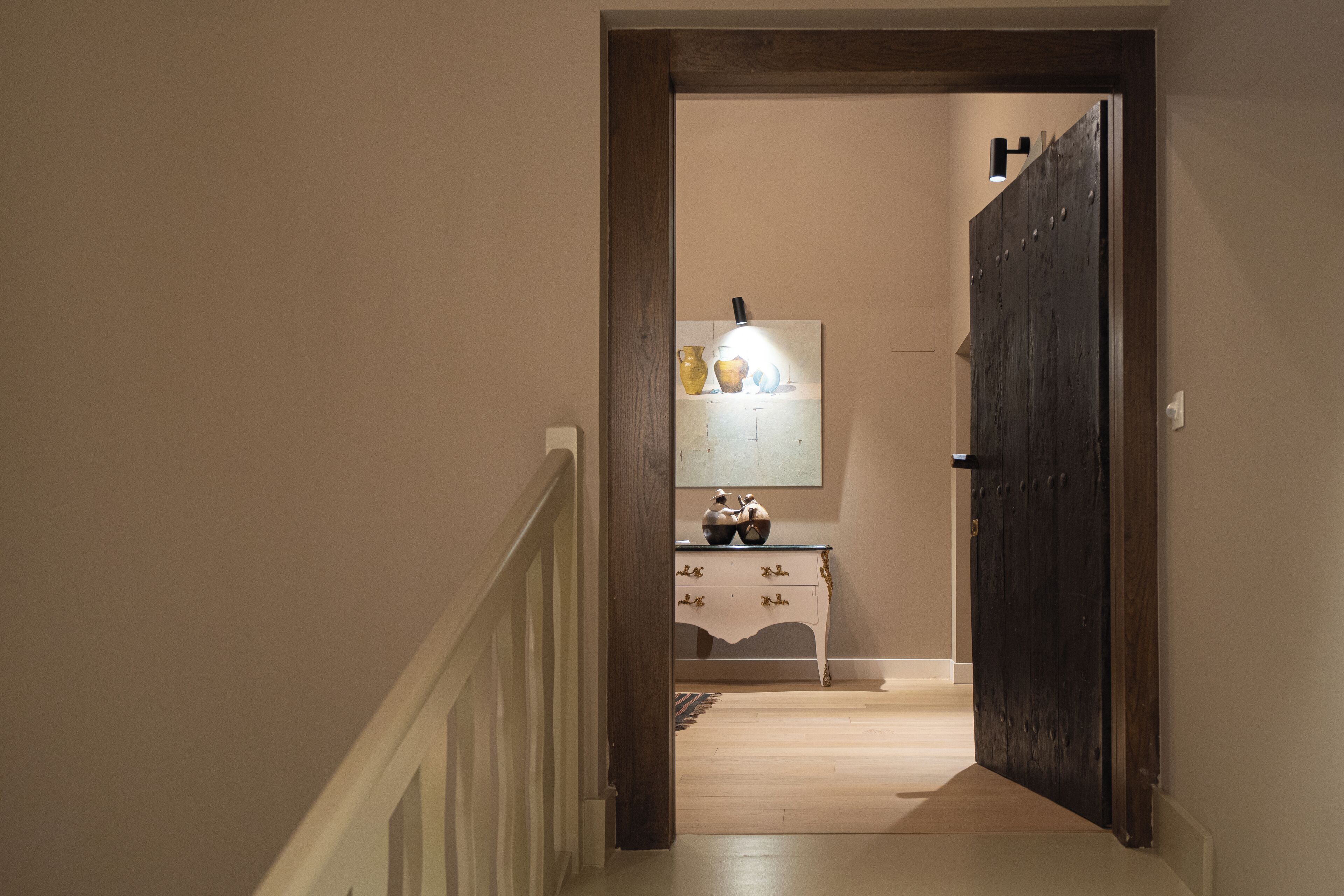 Een gang met een houten deur, een klein tafeltje met decoratieve objecten en een wandlamp.