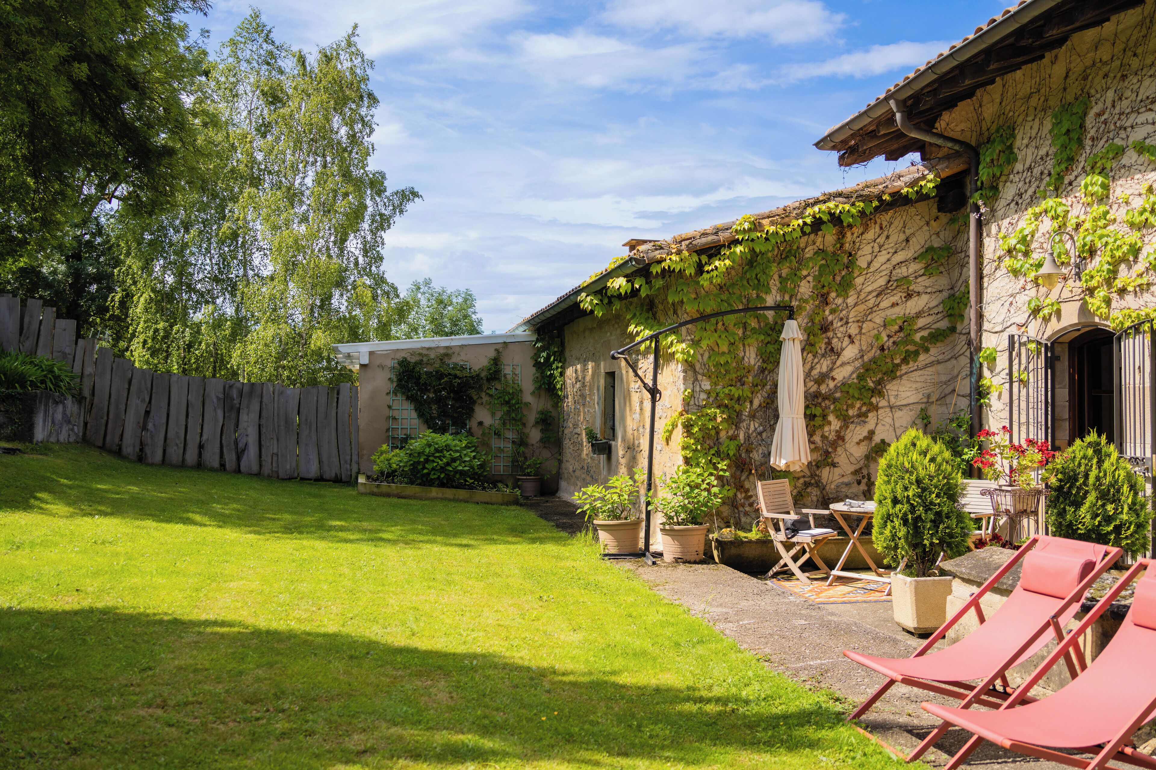 Un edificio in pietra con tetto in tegole, recinzione in legno e giardino con prato. Si notano piante in vaso e mobili da esterno.