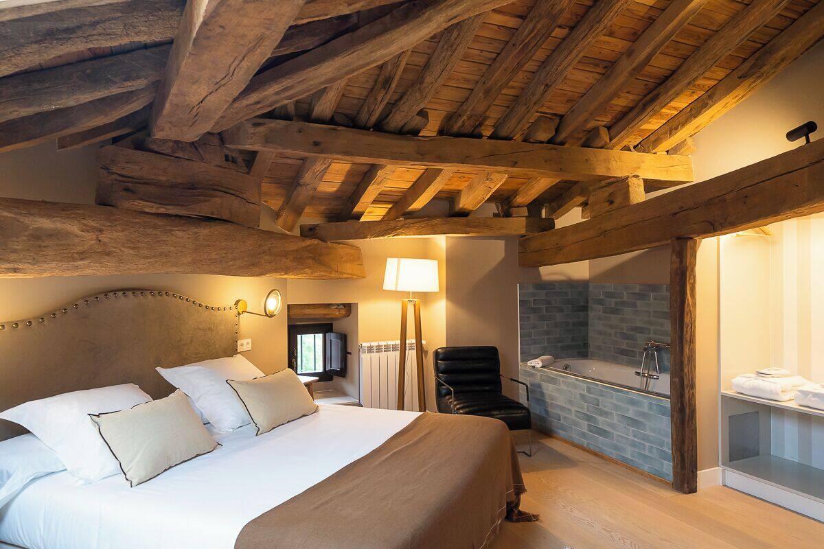 Une chambre avec un plafond en bois, un grand lit avec du linge de lit blanc, un fauteuil en cuir noir, une baignoire intégrée et une petite salle de bain avec un miroir et un porte-serviettes.