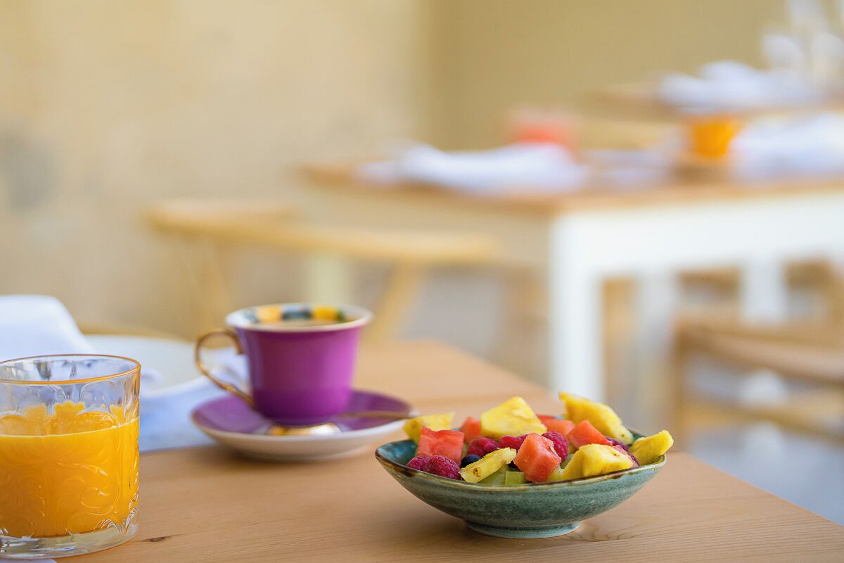 Une table de petit-déjeuner avec un saladier de fruits frais, un verre de jus d’orange et une tasse violette.