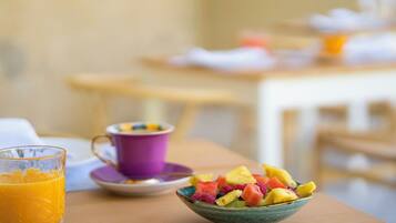 Une table de petit-déjeuner avec un saladier de fruits frais, un verre de jus d’orange et une tasse violette.
