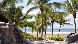 Zona de piscina con palmeras, cascada y vista a la playa.