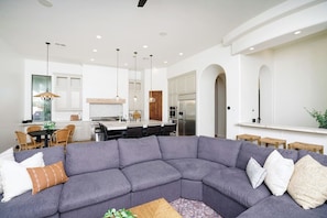 A modern living room with a gray sectional sofa, a dining area with a wooden table and chairs, and a kitchen with white cabinetry and stainless steel appliances.