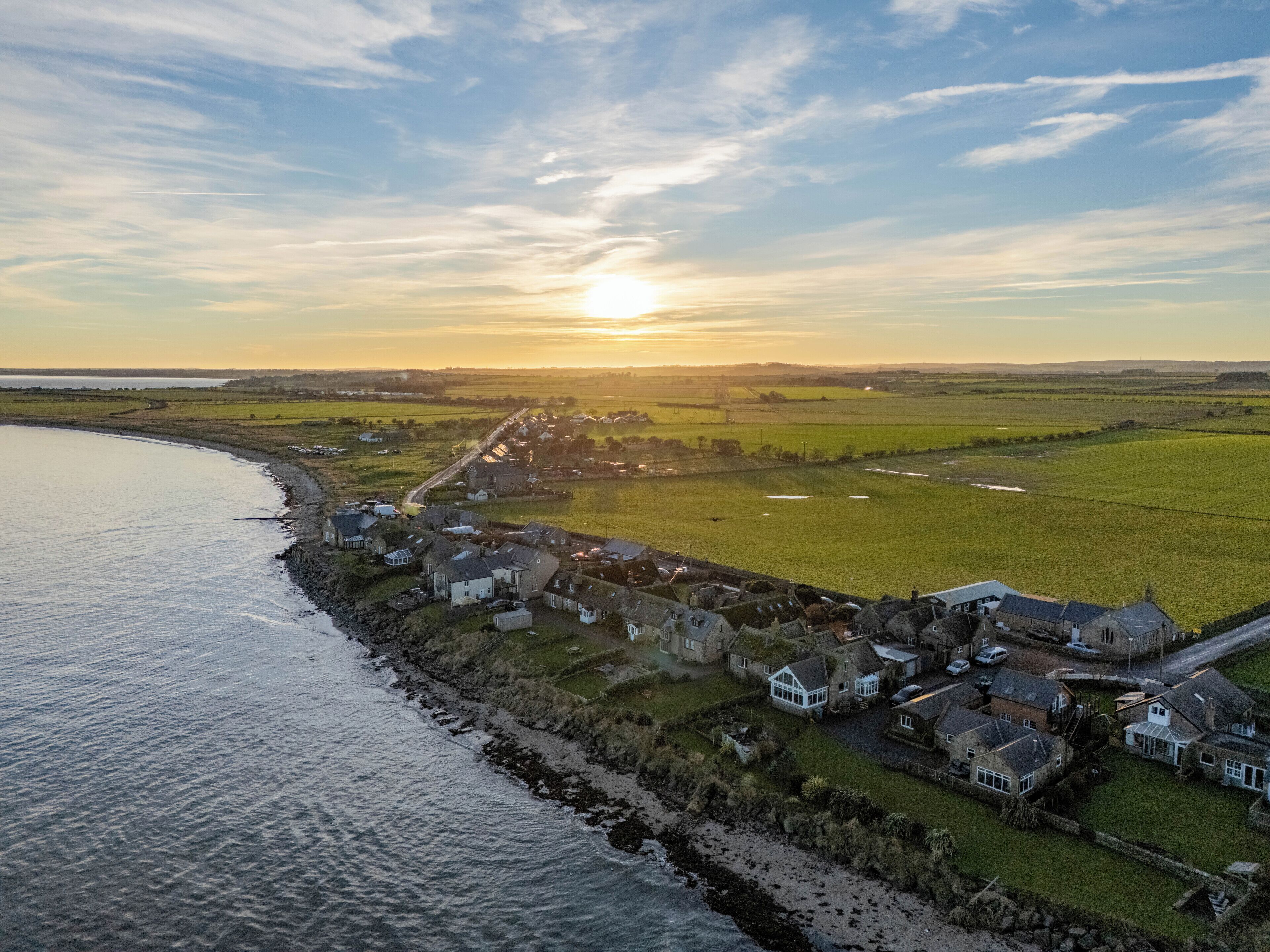 An aerial view of a coastal village with houses along the shoreline, a road leading inland, and fields in the background.