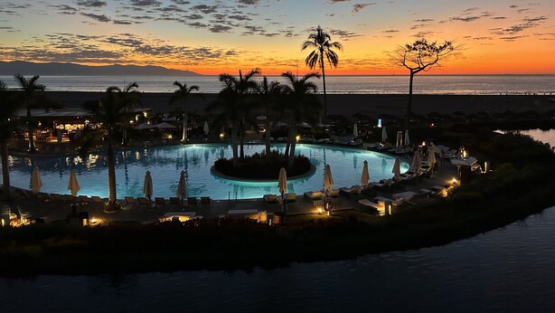 A resort pool with lounge chairs and umbrellas, surrounded by palm trees, with a sunset view.