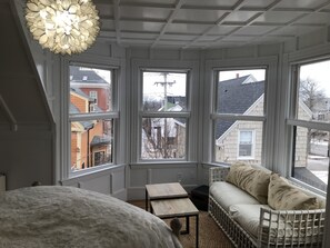 Une chambre avec un grand lit, une table de chevet en bois, un canapé blanc orné de coussins à motifs et une grande fenêtre à carreaux.