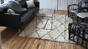 Un salon moderne avec un canapé noir, une table basse en verre, un tapis à motifs et un luminaire suspendu hexagonal original.