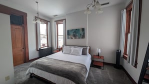 Une chambre à coucher avec un lit, une table de chevet, une lampe, un tableau et une fenêtre avec des rideaux.