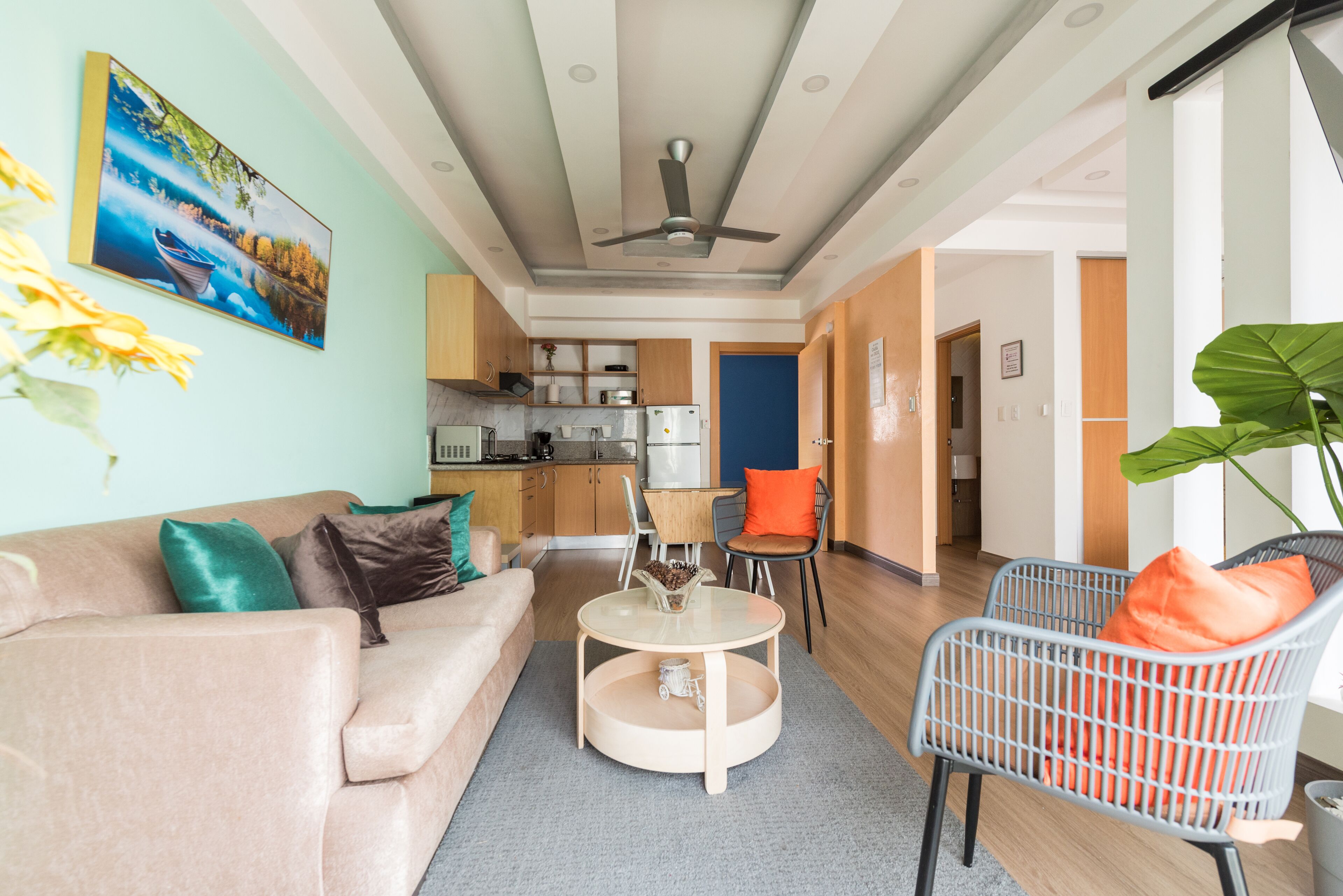A modern living room with a sofa, coffee table, and chairs, a kitchen in the background, and a ceiling fan.