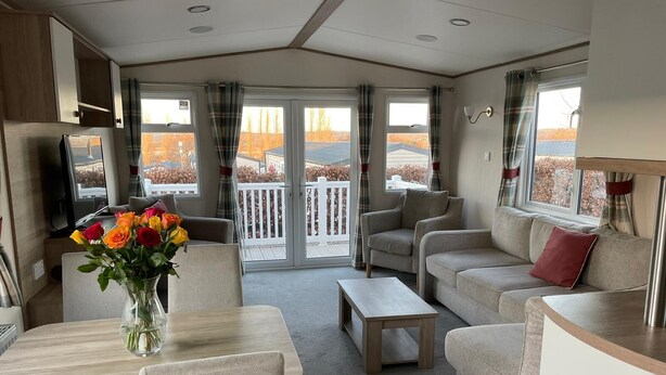 A modern, well-lit living area with a sofa, armchairs, a coffee table, and a dining table. A vase of flowers is on the table. Large windows offer a view of a deck and trees. - Arum dog friendly caravan on Rockley Park, Poole (Poole)