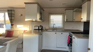 A compact kitchen with white cabinets, a stove, and a sink. There is a window with blinds and a view of the outside. - Arum dog friendly caravan on Rockley Park, Poole (Poole)
