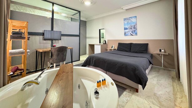 A modern hotel room with a large bed, a wooden chair, a desk, a TV, a painting, and a bathtub with toiletries. - Grond Konaklama Mersin (Mersin)