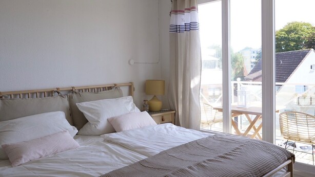 A bedroom with a bed, a bedside lamp, a window with curtains, and a view of buildings outside.