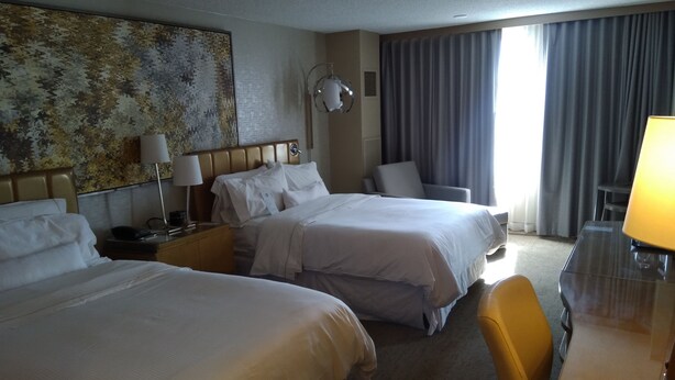 Une chambre d’hôtel avec deux lits, un bureau, une chaise, une lampe et une grande fenêtre avec des rideaux.