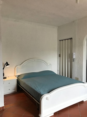 Une chambre avec un lit simple, une tête de lit blanche, une table de chevet avec une lampe et une porte équipée de stores verticaux.