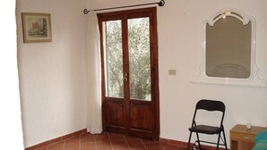 A room with a wooden door, a window with a view of trees, a chair, and a small table with a lamp. - Four - Miguel Apartment with panoramic view, among olive trees, sea 15 km away (Villa Viani)