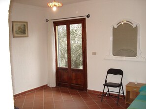 A room with a wooden door, a window with a view of trees, a chair, and a small table with a lamp.