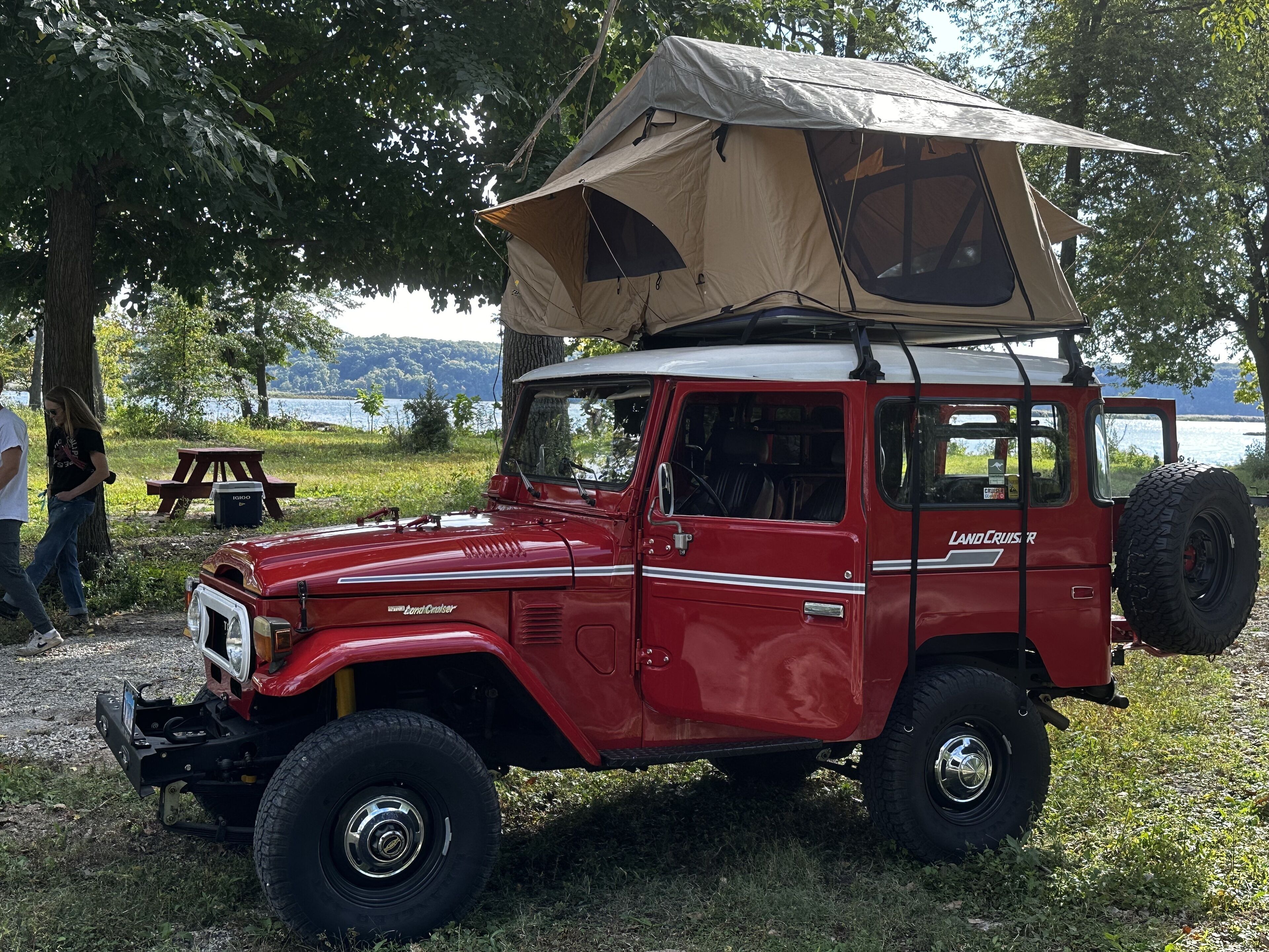 Ein roter Land Cruiser mit einer Lienwandverdeck, geparkt in der Nähe eines Picknicktisches und eines Sees.