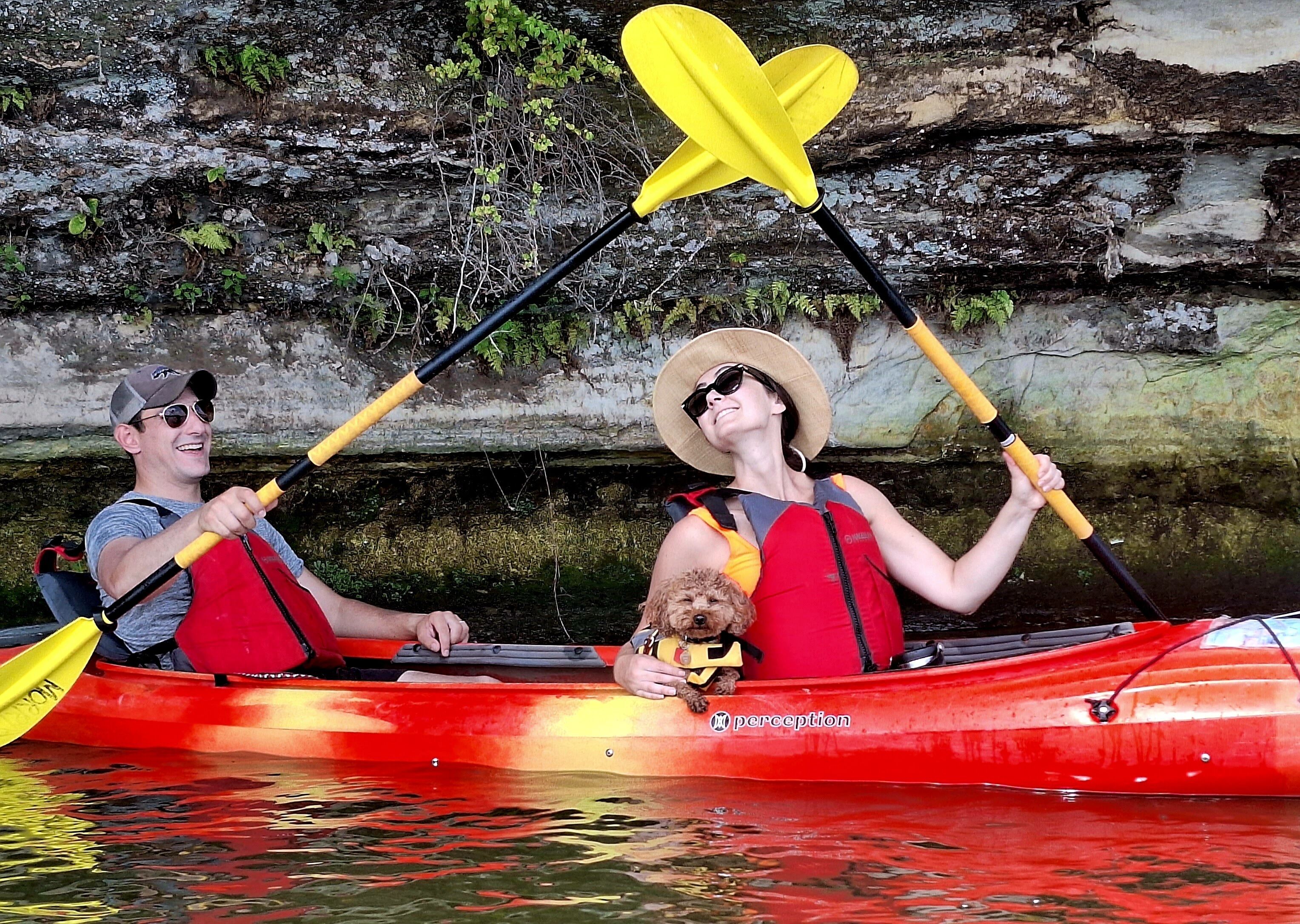 Two people kayaking in a red canoe with a dog, using yellow paddles, and wearing life jackets.