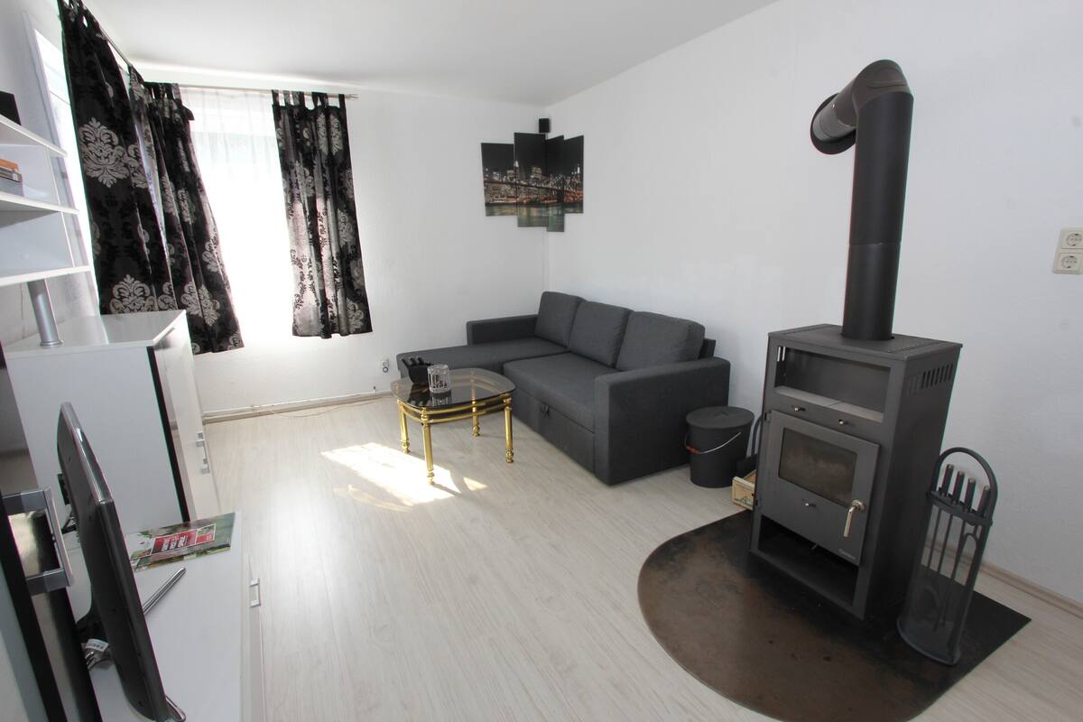 Een moderne woonkamer met een grijze bank, een klein bijzettafeltje, een zwarte houtkachel en een raam met zwarte gordijnen.