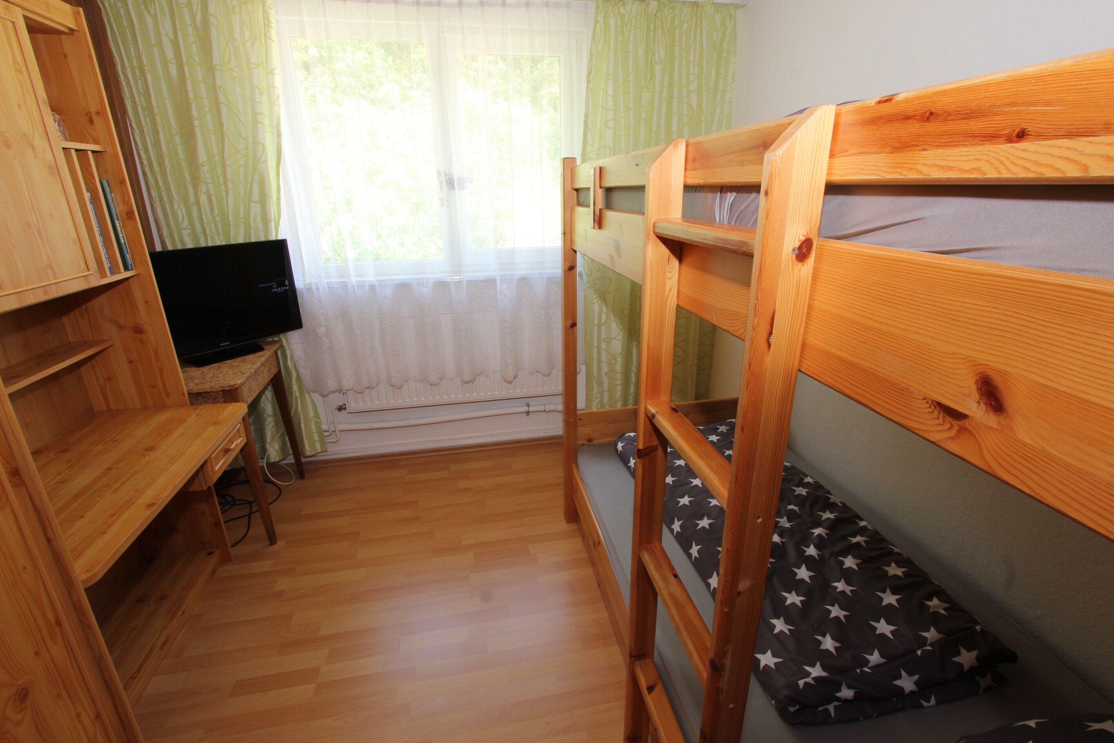 Une chambre avec un lit superposé, des meubles en bois, une télévision et une fenêtre ornée de rideaux légers.