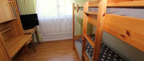 Une chambre avec un lit superposé, des meubles en bois, une télévision et une fenêtre ornée de rideaux légers.