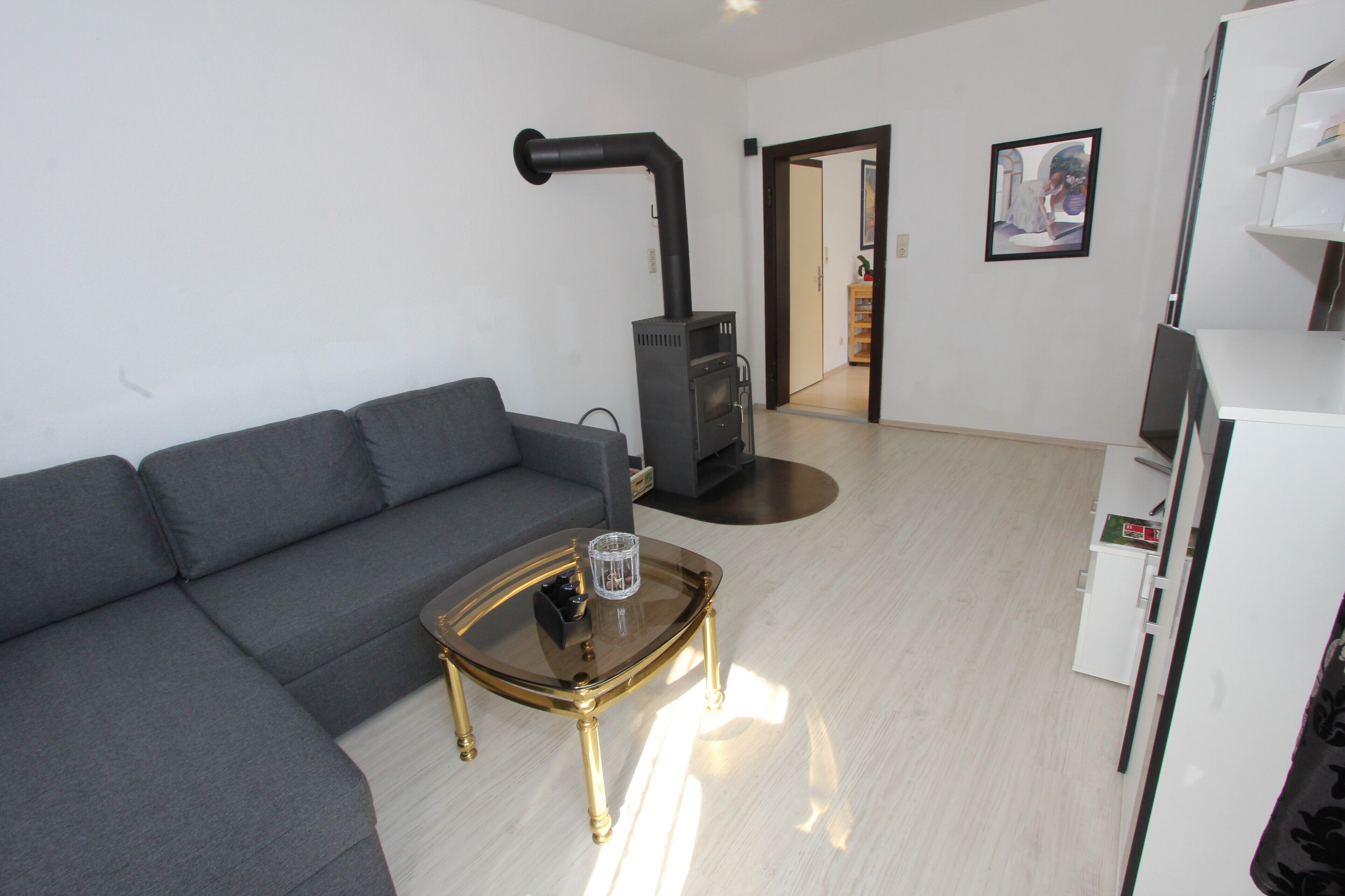 Een woonkamer met een grijze bank, een glazen salontafel met gouden poten, een zwarte verwarming en een witte koelkast.