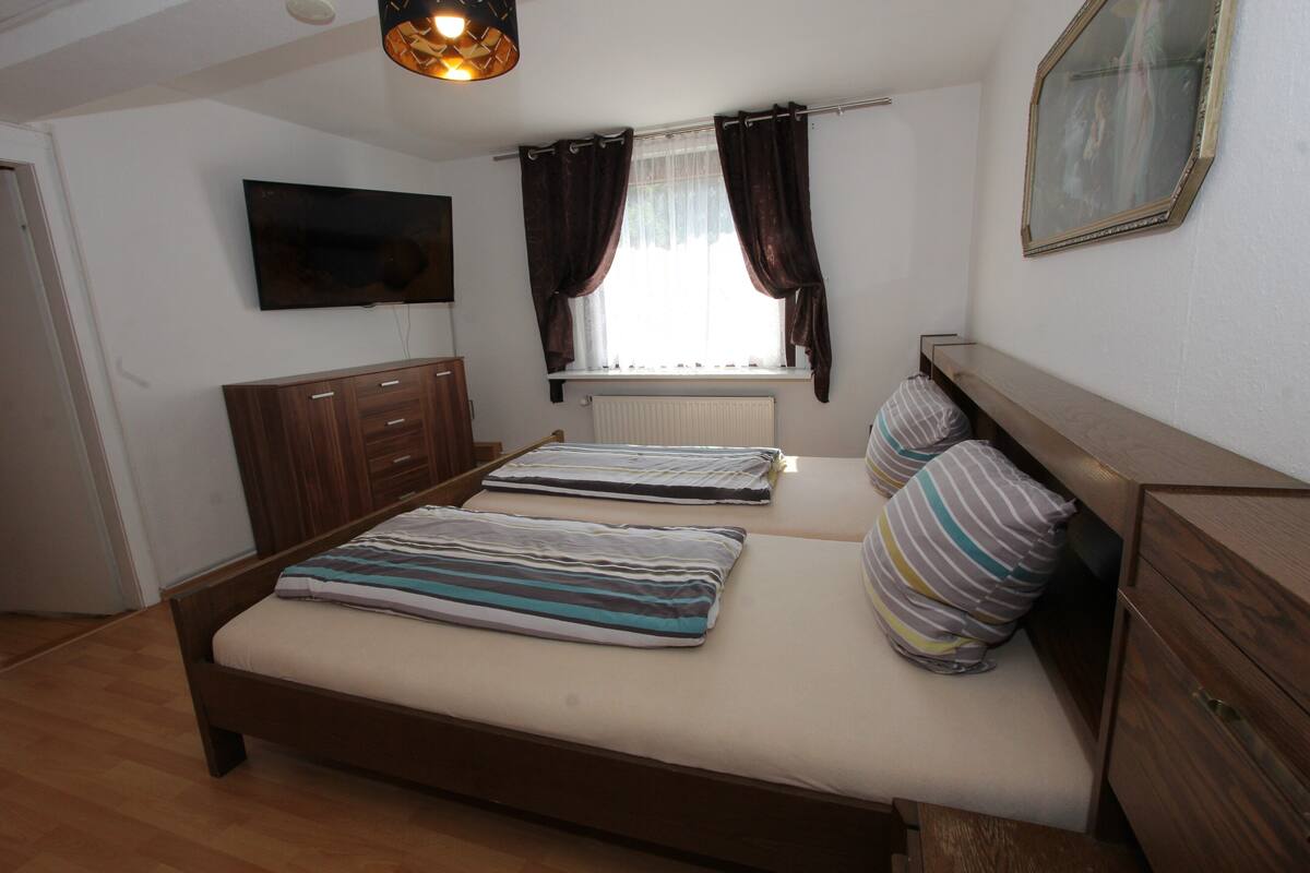 Een slaapkamer met twee bedden, een televisie, een raam met gordijnen, een radiator en een houten dressoir.