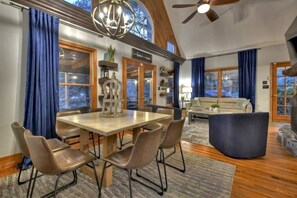 A spacious living room with a high ceiling, a dining area with a wooden table and chairs, and a cozy seating area with a sofa and ottoman.