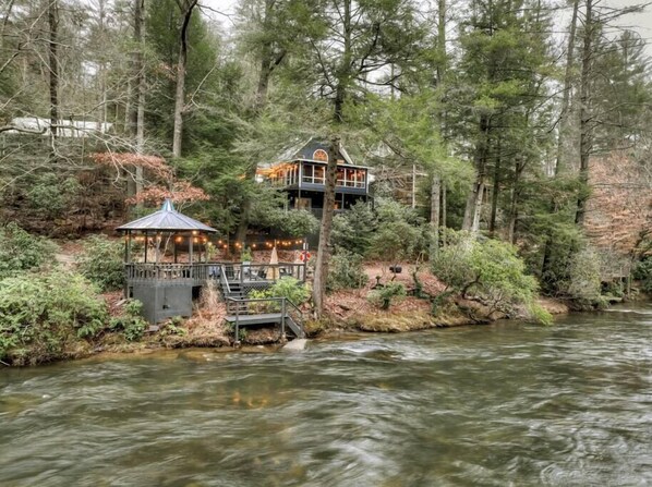 A cabin-style house with a deck and a gazebo by a river, surrounded by dense forest.