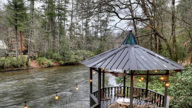 Ein Pavillon mit rundem Tisch und Stühlen, mit Blick auf einen Fluss, umgeben von Bäumen.