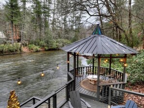 A gazebo with a circular table and chairs, overlooking a river, surrounded by trees.