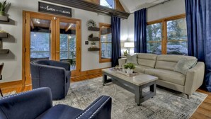 Ein gemütliches Wohnzimmer mit einer Couch, zwei Sesseln, einem Couchtisch und einem Kamin. Die Decke ziert ein Holzbalkenbau, und ein großes Fenster ist mit Vorhängen versehen.