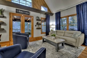 A cozy living room with a sofa, two armchairs, a coffee table, and a fireplace. There are wooden beams on the ceiling and a large window with curtains. - Riverside Hideaway | $229 Off Your Stay! (Blue Ridge)