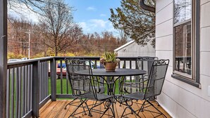 Eine Holzterrasse mit einem Metalltisch und -stühlen, einem Blumentopf und Blick auf einen Parkplatz sowie Bäume.