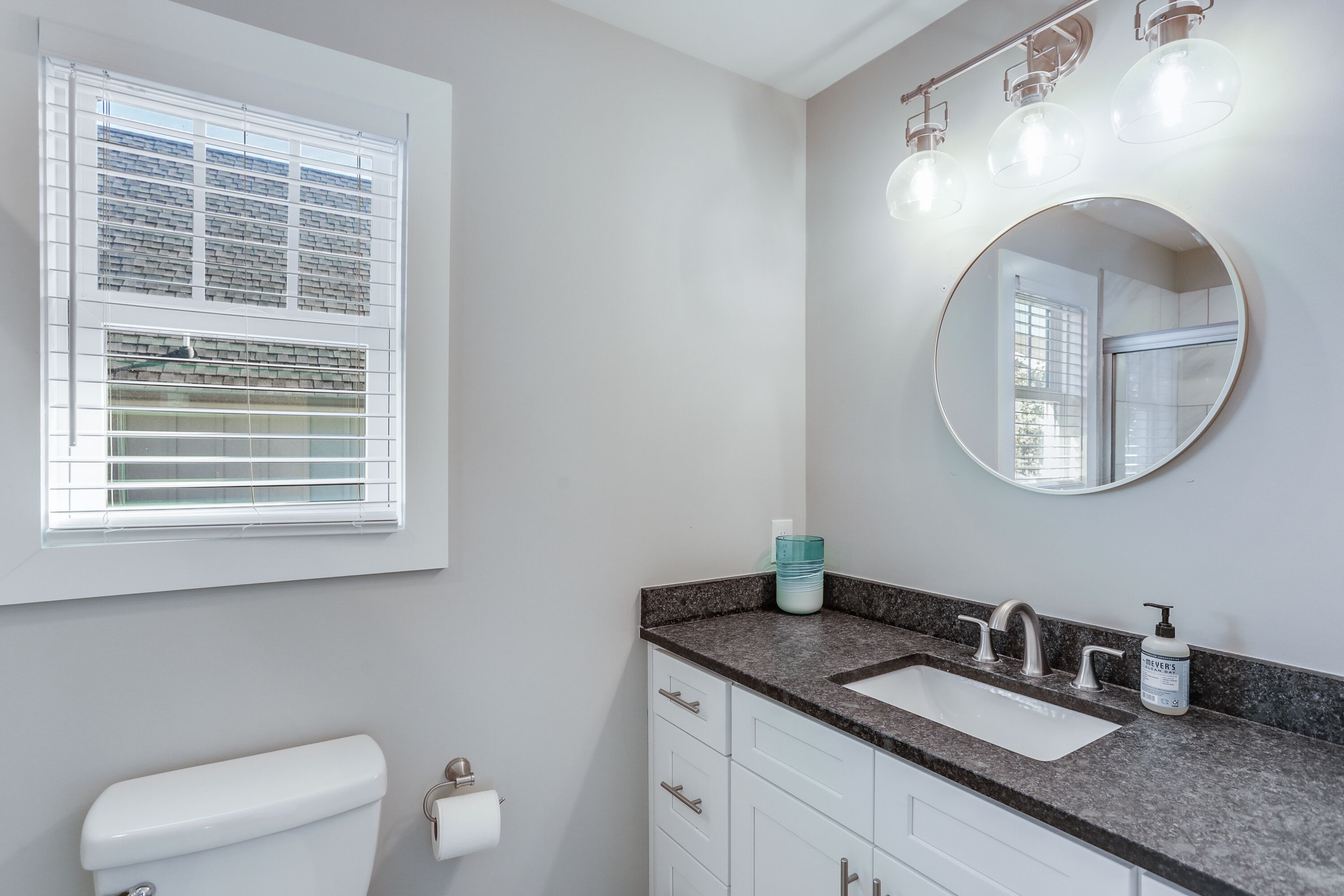 Une salle de bain avec des toilettes, un lavabo et un miroir. Il y a une fenêtre avec des stores et un flacon de savon liquide sur le comptoir du lavabo.