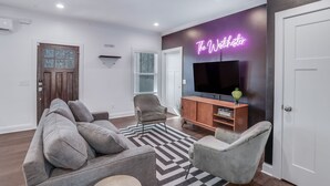 Un salon moderne avec un canapé gris, des fauteuils, un téléviseur et un panneau lumineux néon.