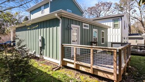 Une maison à deux étages avec une façade verte, une terrasse en bois dotée d’une rambarde en métal et un petit porche.