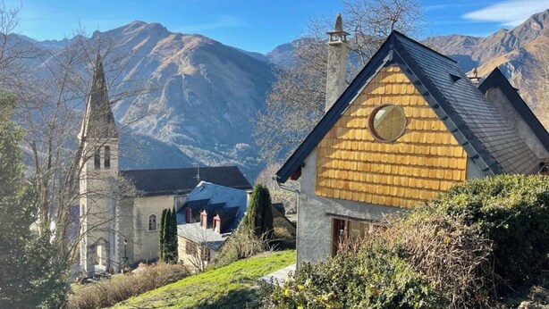 A traditional wooden house with a stone church in the background, surrounded by mountains.