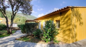Un bâtiment jaune avec un toit en tuiles, entouré de verdure et bordé d’un sentier.