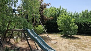 Un parque infantil con tobogán y columpios, rodeado de árboles y suelo de tierra.