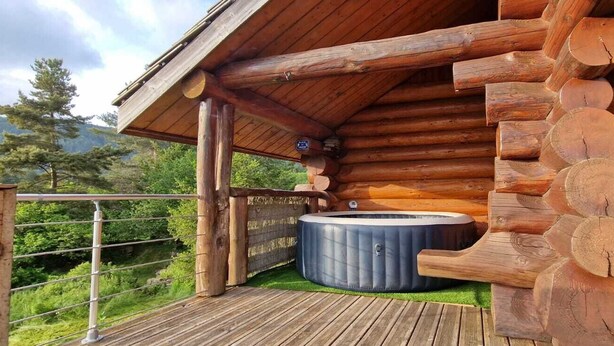 Una cabaña de troncos con un jacuzzi en una terraza de madera, rodeada de follaje.
