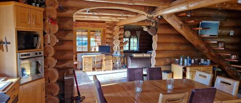 A log cabin interior with a kitchen, dining area, and a large wooden dining table with chairs.