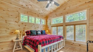 Un dormitorio de cabaña de madera con una cama, dos ventanas y un ventilador de techo.