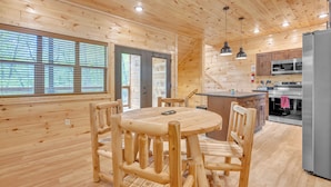 Interior de una cabaña de madera con una mesa de comedor, sillas, una zona de cocina con refrigerador, microondas y estufa.