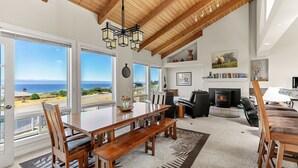 A spacious living room with a wooden dining table, benches, and a fireplace. Large windows offer a view of the ocean. - Stunning views near beaches and bluff (Sea Ranch)